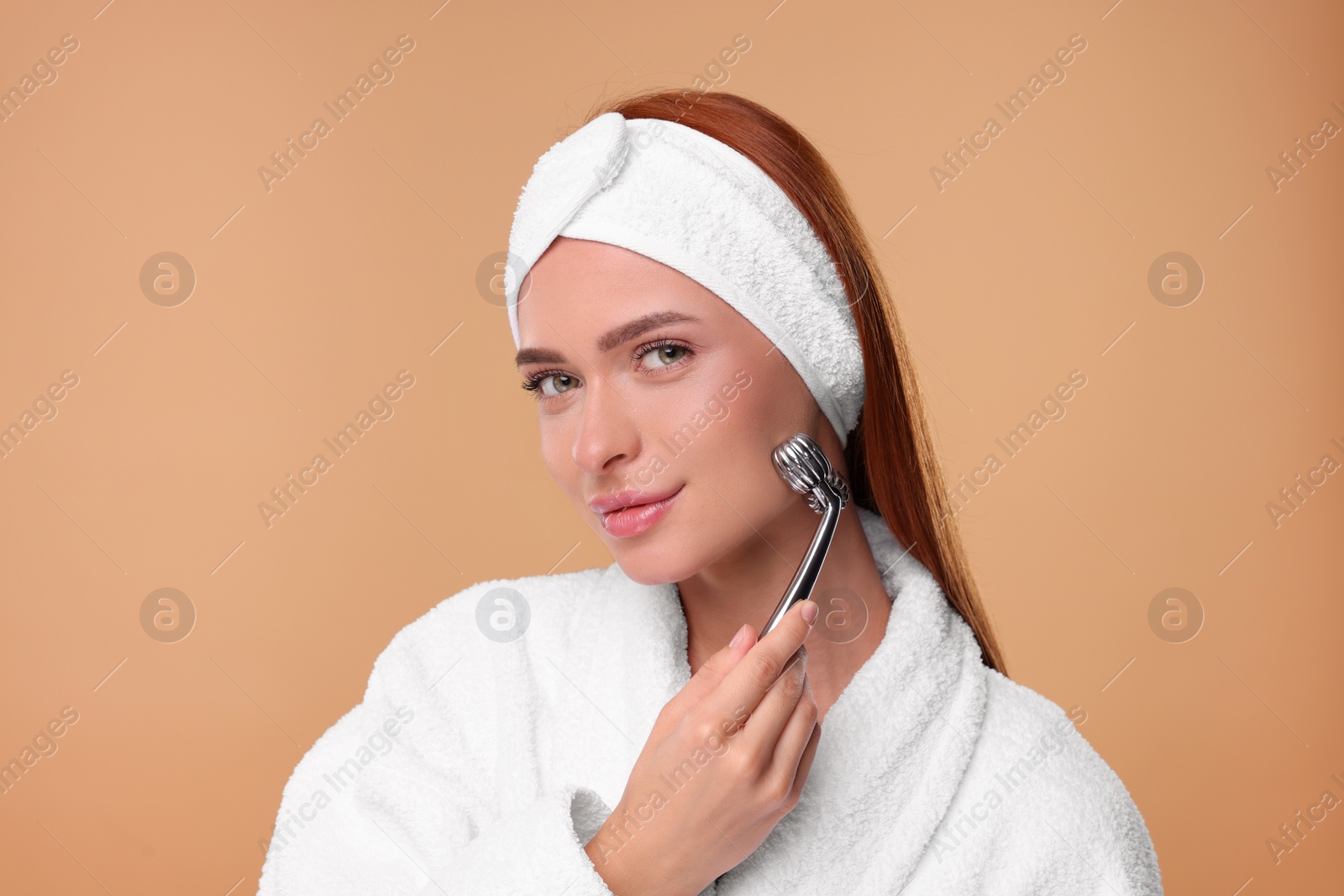 Photo of Young woman massaging her face with metal roller on pale orange background