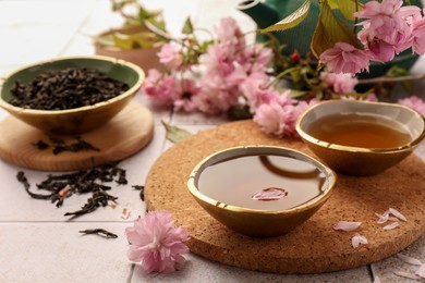 Photo of Traditional ceremony. Cup of brewed tea, teapot, dried leaves and sakura flowers on tiled table, closeup
