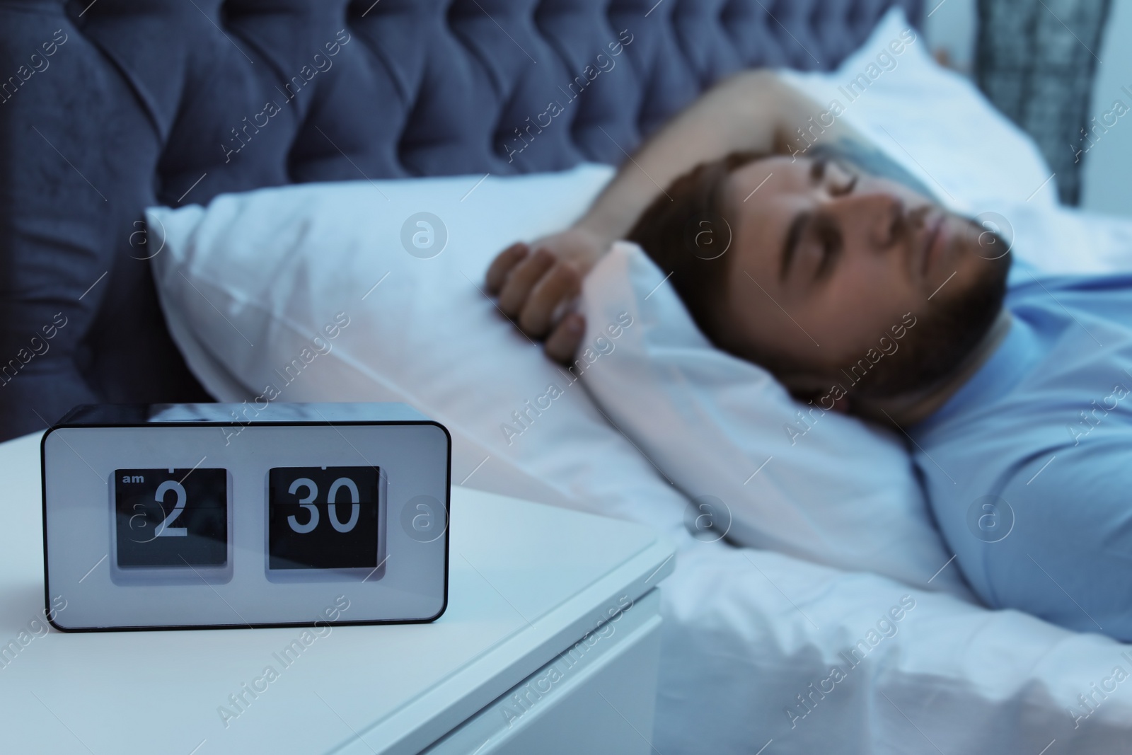 Photo of Alarm clock on table and young man sleeping in bed at night