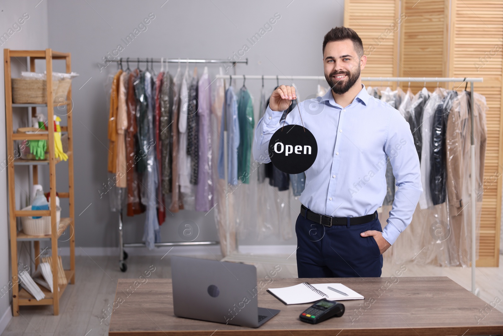 Photo of Dry-cleaning service. Happy worker holding Open sign at workplace indoors, space for text