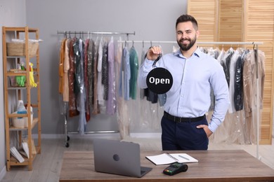 Photo of Dry-cleaning service. Happy worker holding Open sign at workplace indoors, space for text