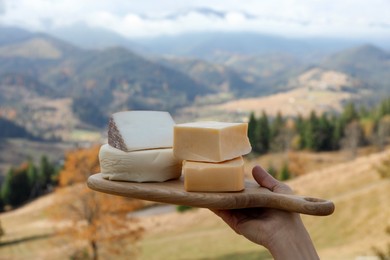 Woman holding board with different types of delicious cheeses against mountain landscape, closeup