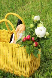 Photo of Yellow wicker bag with wine, bread and flowers on green grass outdoors. Picnic season