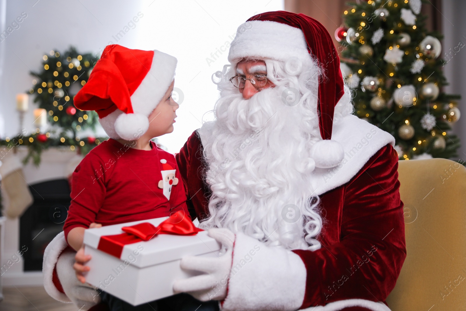 Photo of Santa Claus giving present to little boy in room decorated for Christmas