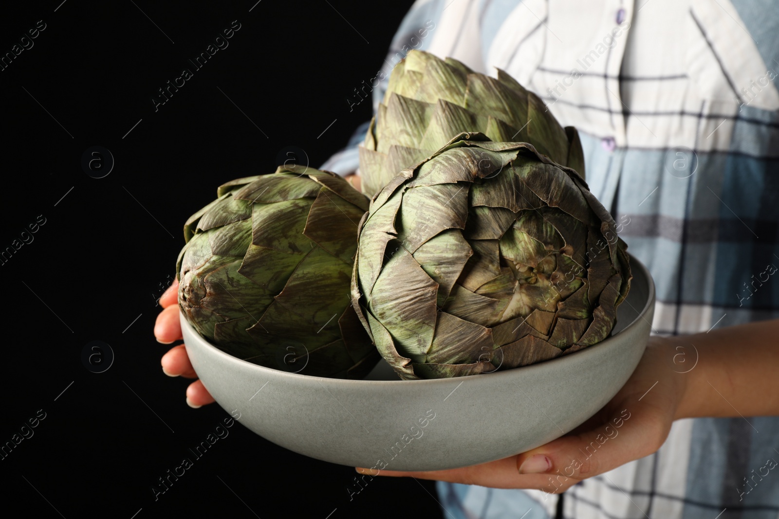 Photo of Woman holding bowl with fresh raw artichokes on black background, closeup