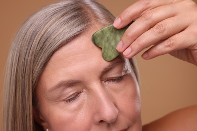 Woman massaging her face with jade gua sha tool on brown background, closeup