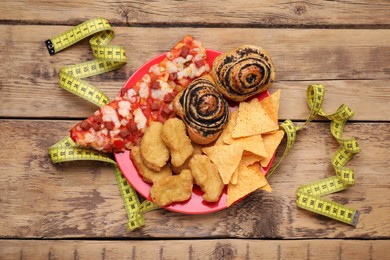 Photo of Plate of different unhealthy food and measuring tape on wooden table, flat lay. Weight loss concept