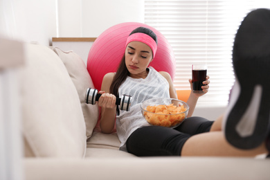 Photo of Lazy young woman with sport equipment and junk food at home