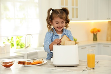 Cute little girl putting slice of bread into toaster at table in kitchen