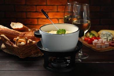 Photo of Fork with piece of broccoli, melted cheese in fondue pot, wine and snacks on wooden table, closeup