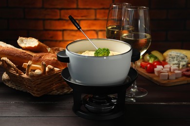 Fork with piece of broccoli, melted cheese in fondue pot, wine and snacks on wooden table, closeup