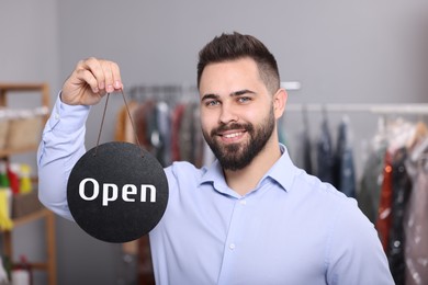 Photo of Dry-cleaning service. Happy worker holding Open sign indoors