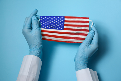 Doctor holding medical mask with USA flag pattern on light blue background, closeup. Dangerous virus