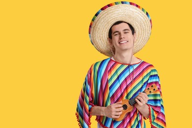 Photo of Young man in Mexican sombrero hat and poncho playing ukulele on yellow background. Space for text