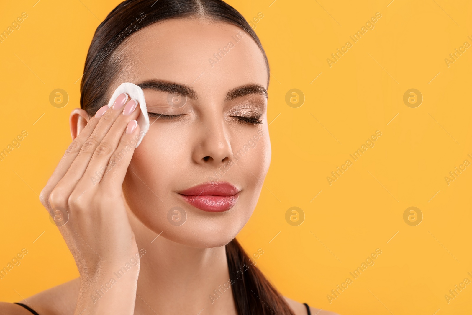 Photo of Beautiful woman removing makeup with cotton pad on orange background, space for text