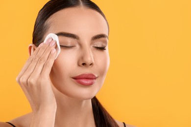 Beautiful woman removing makeup with cotton pad on orange background, space for text