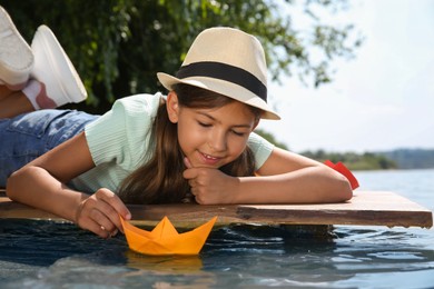 Cute little girl playing with paper boat on wooden pier near river