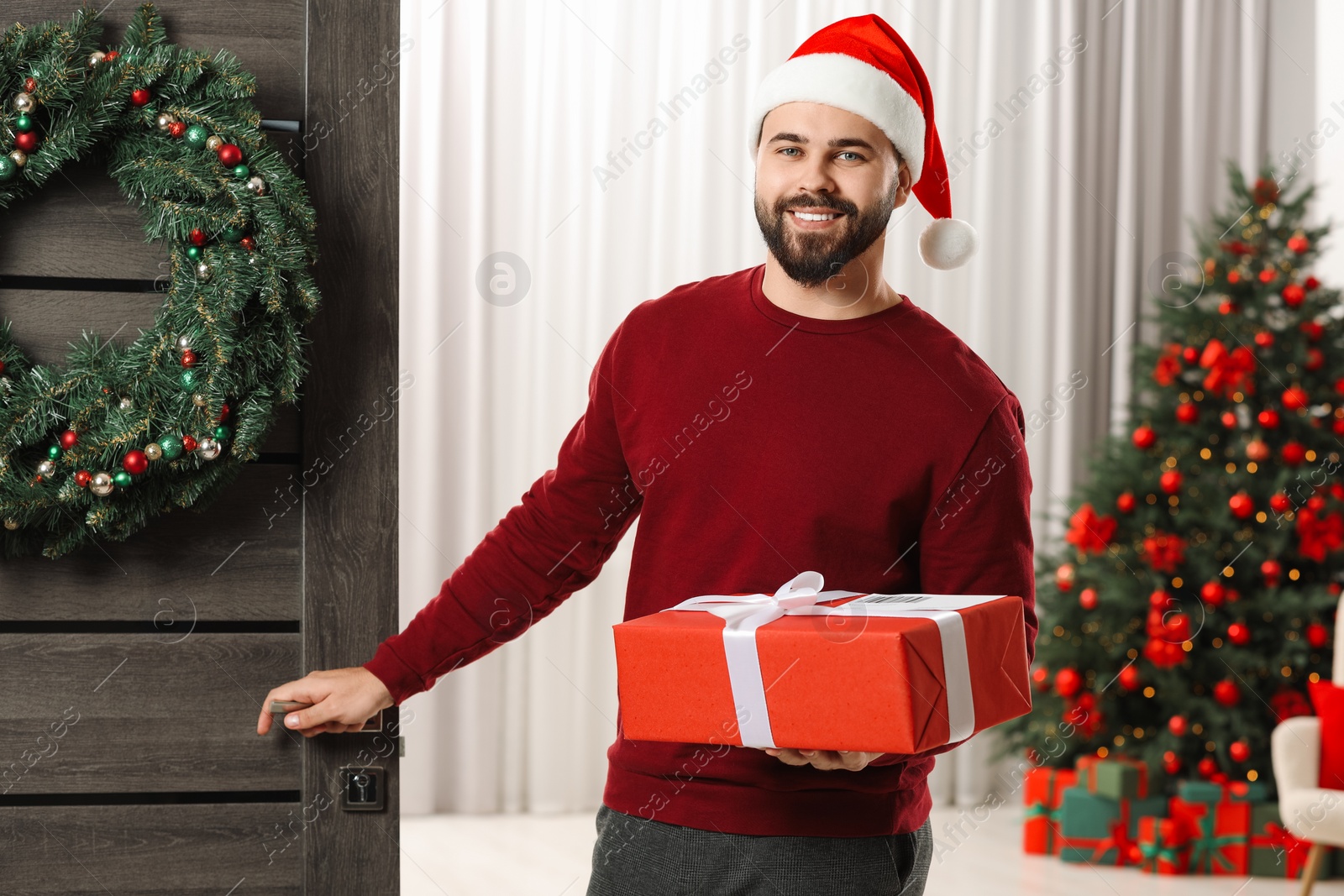 Photo of Young man in Santa hat with Christmas gift box received by mail indoors