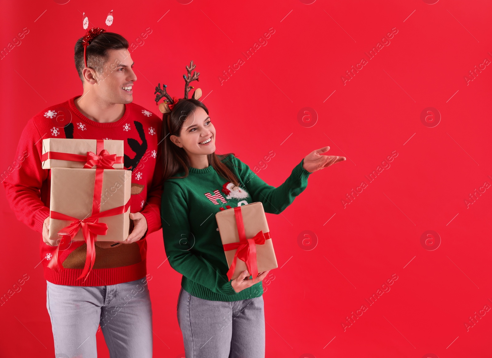 Photo of Beautiful happy couple in Christmas headbands and sweaters holding gifts on red background. Space for text