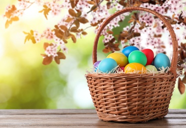 Image of Colorful Easter eggs in wicker basket on wooden table outdoors, space for text