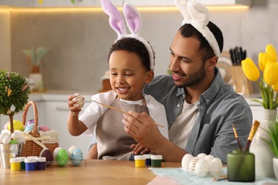 Happy African American father and his cute son painting Easter eggs at table in kitchen
