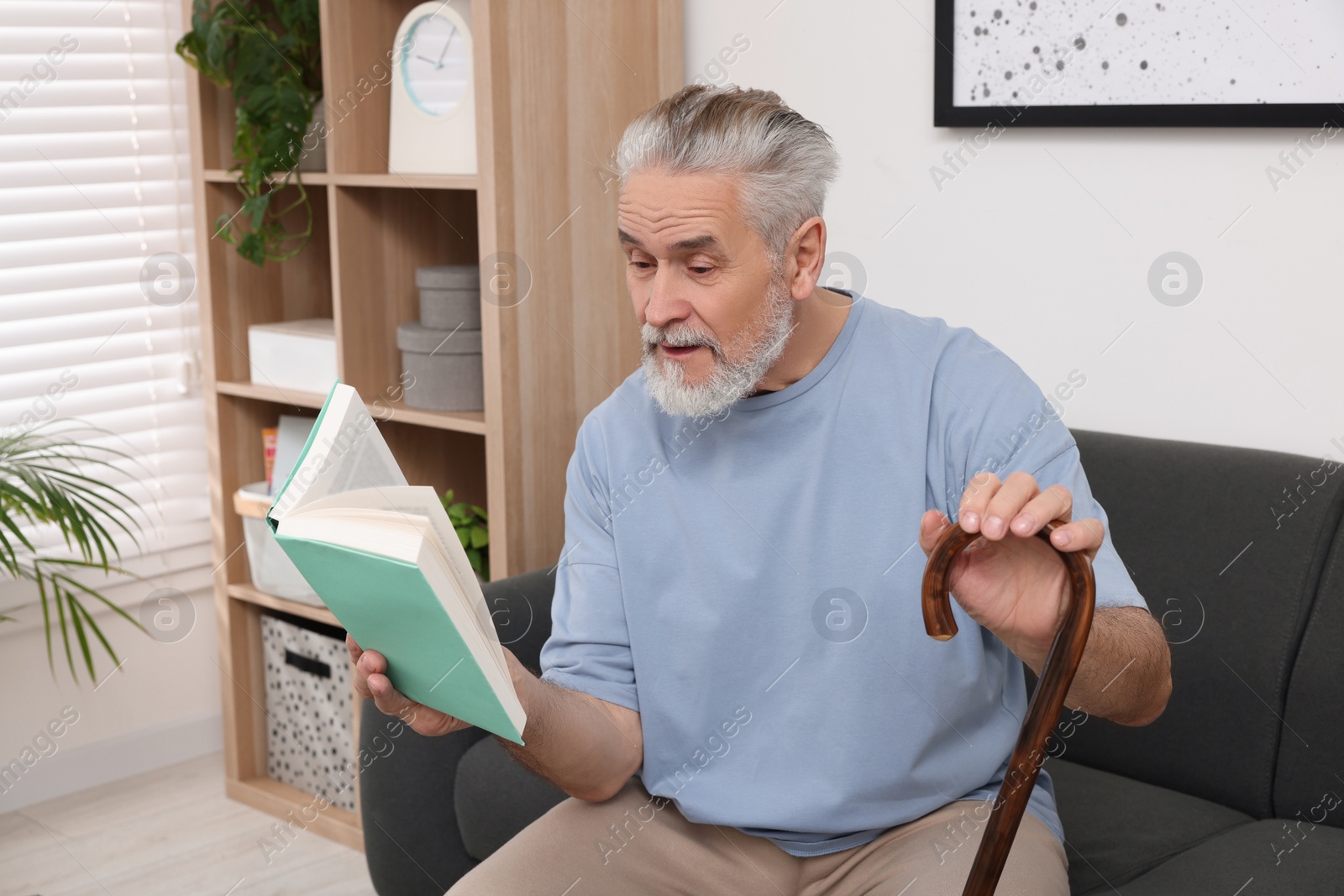 Photo of Senior man with walking cane reading book on sofa at home