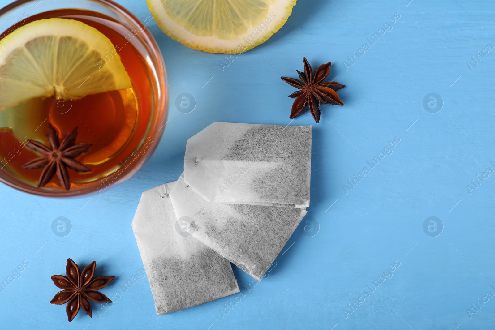 Photo of Tea bags, cup of hot drink, anise stars and lemon on light blue wooden table, flat lay. Space for text