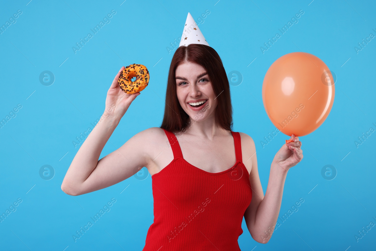 Photo of Happy woman in party hat with doughnut and balloon on light blue background