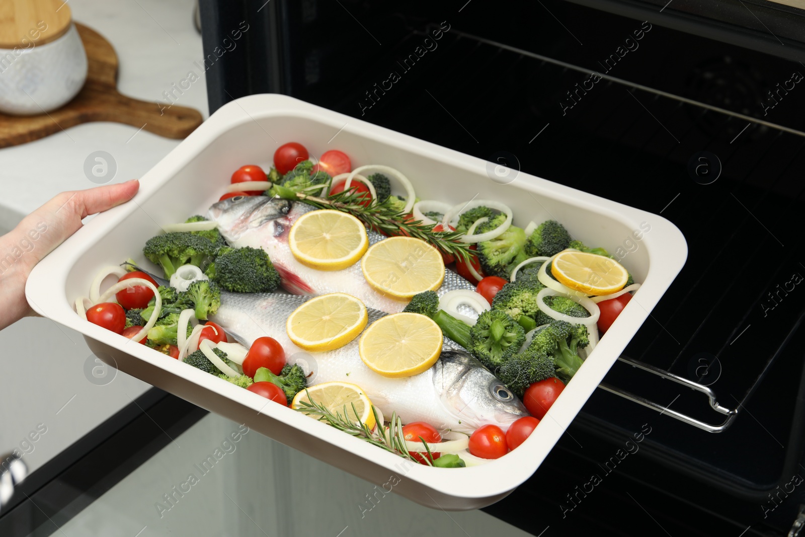 Photo of Woman putting baking dish with raw fish and vegetables into oven in kitchen, closeup