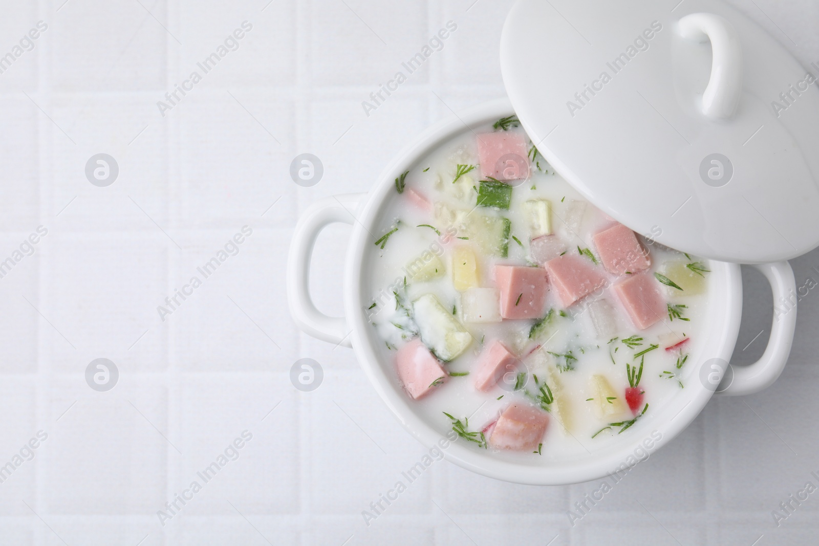 Photo of Delicious cold summer soup (okroshka) with boiled sausage in pot on white tiled table, top view. Space for text