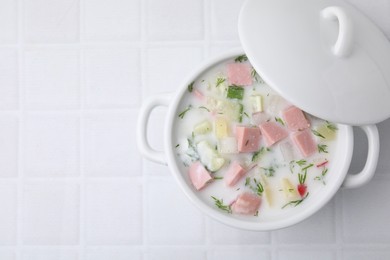 Photo of Delicious cold summer soup (okroshka) with boiled sausage in pot on white tiled table, top view. Space for text