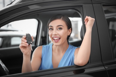 Young woman with key sitting in driver's seat of new car at salon