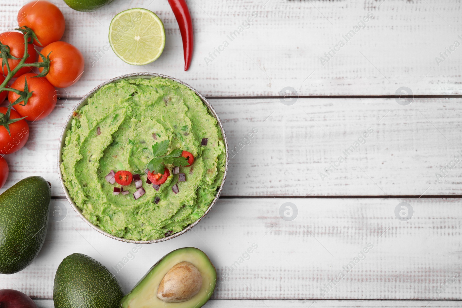Photo of Bowl of delicious guacamole and ingredients on white wooden table, flat lay. Space for text