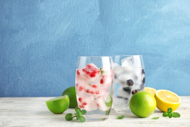 Glasses of drinks with fruit ice cubes on table against color background