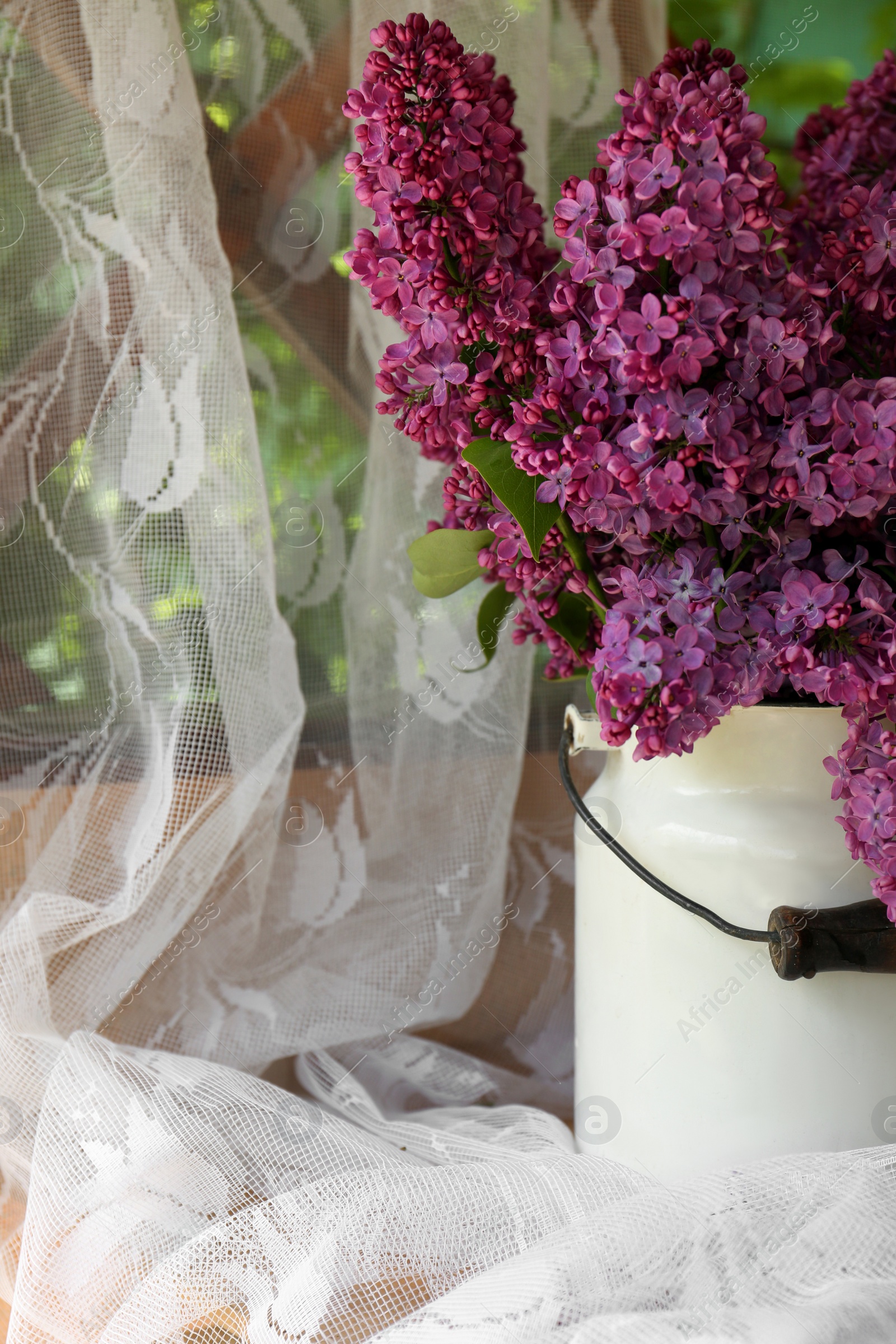 Photo of Beautiful lilac flowers in milk can near window indoors, closeup
