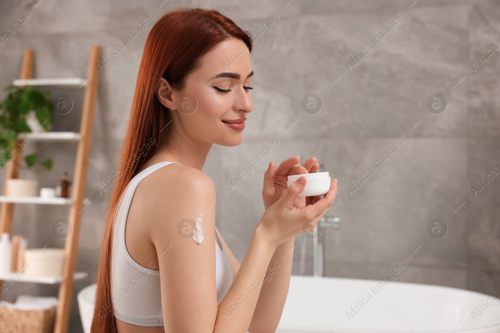 Photo of Beautiful young woman applying body cream onto shoulder in bathroom, space for text