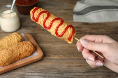 Photo of Woman holding delicious corn dog with ketchup at wooden table, closeup