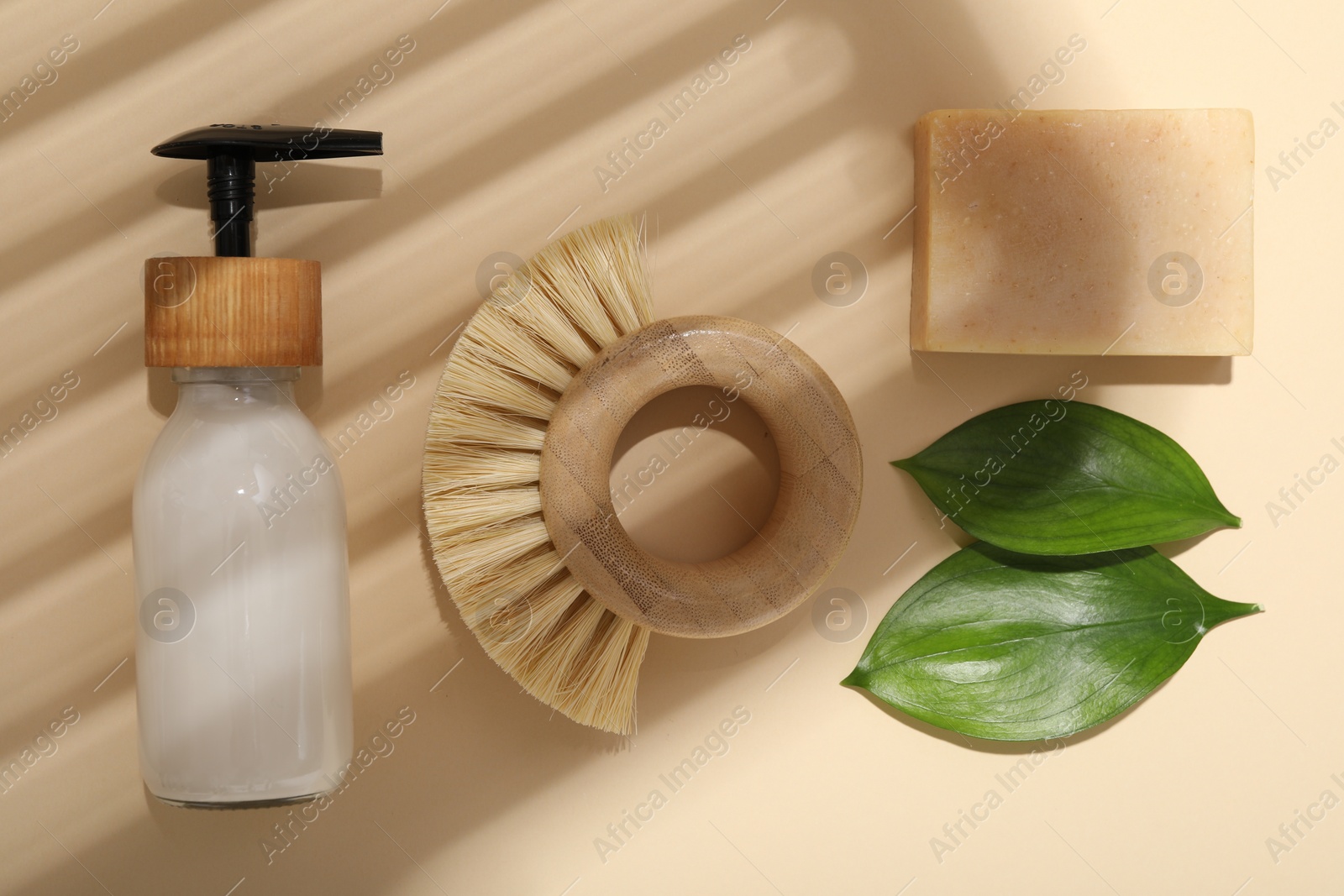 Photo of Cleaning brush, soap bar, dispenser and green leaves on beige background, flat lay