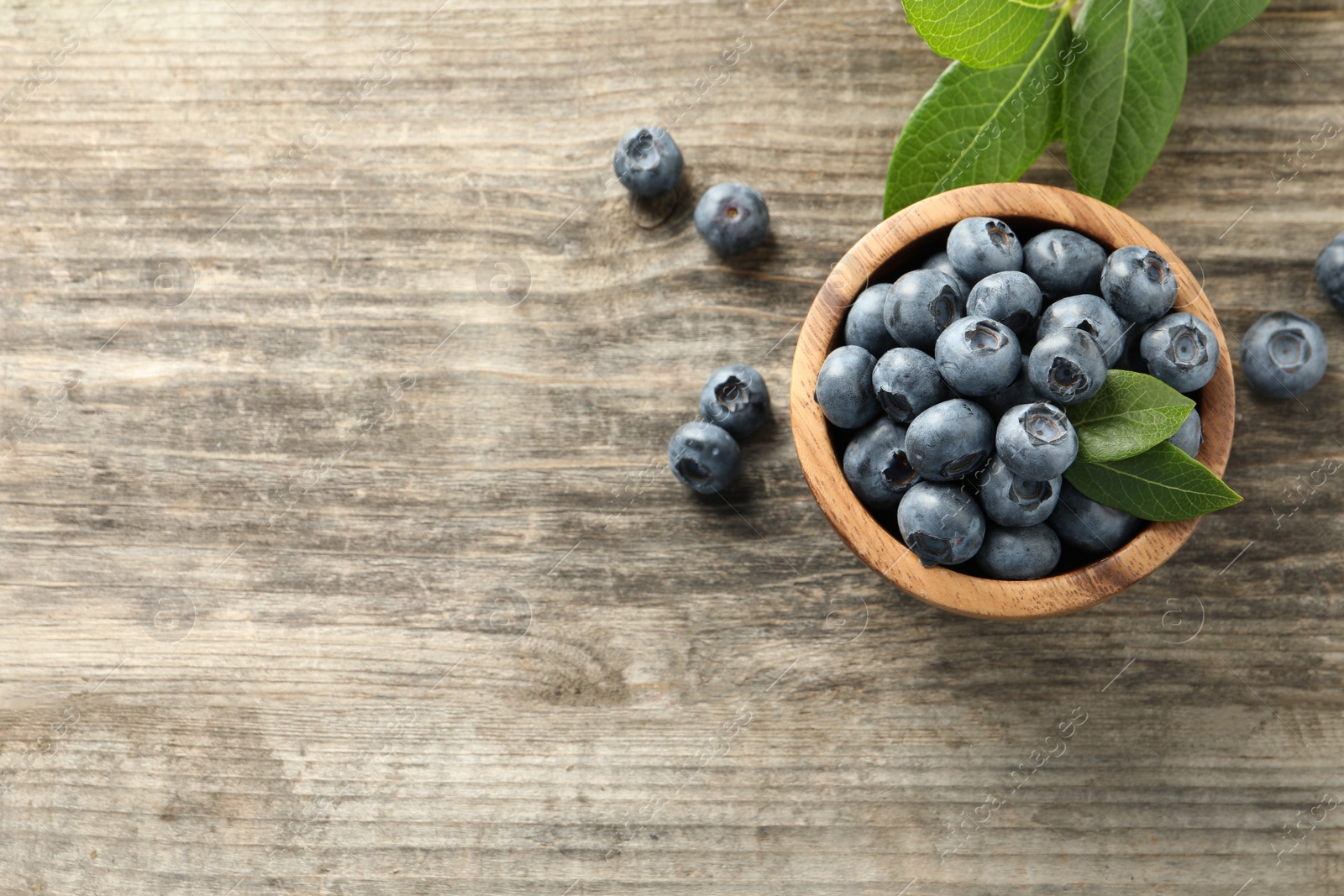 Photo of Bowl of tasty fresh blueberries with leaves on wooden table, flat lay. Space for text