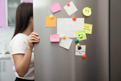 Photo of Woman opening refrigerator door with paper sheets and magnets at home, closeup