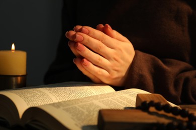 Woman praying at table with burning candle and Bible, closeup