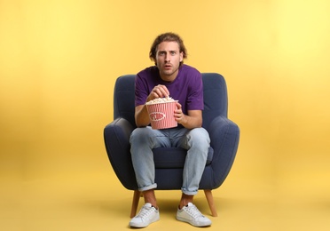 Emotional man with popcorn sitting in armchair during cinema show on color background