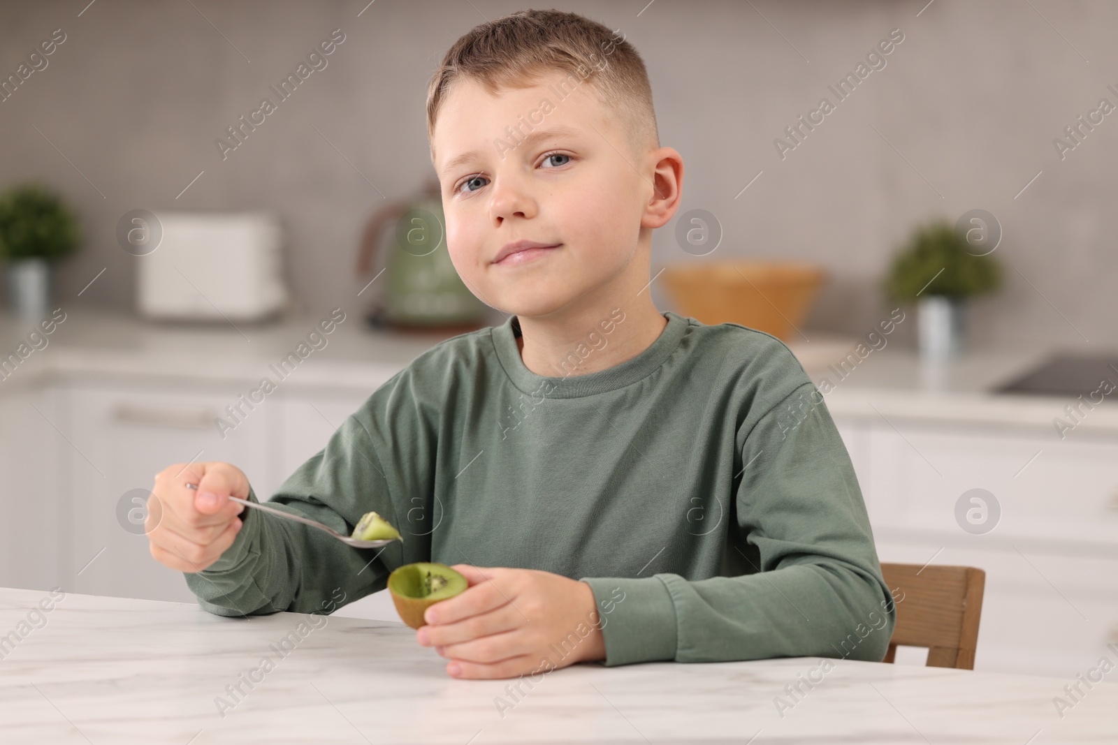 Photo of Boy with tasty fresh kiwi at white marble table in kitchen