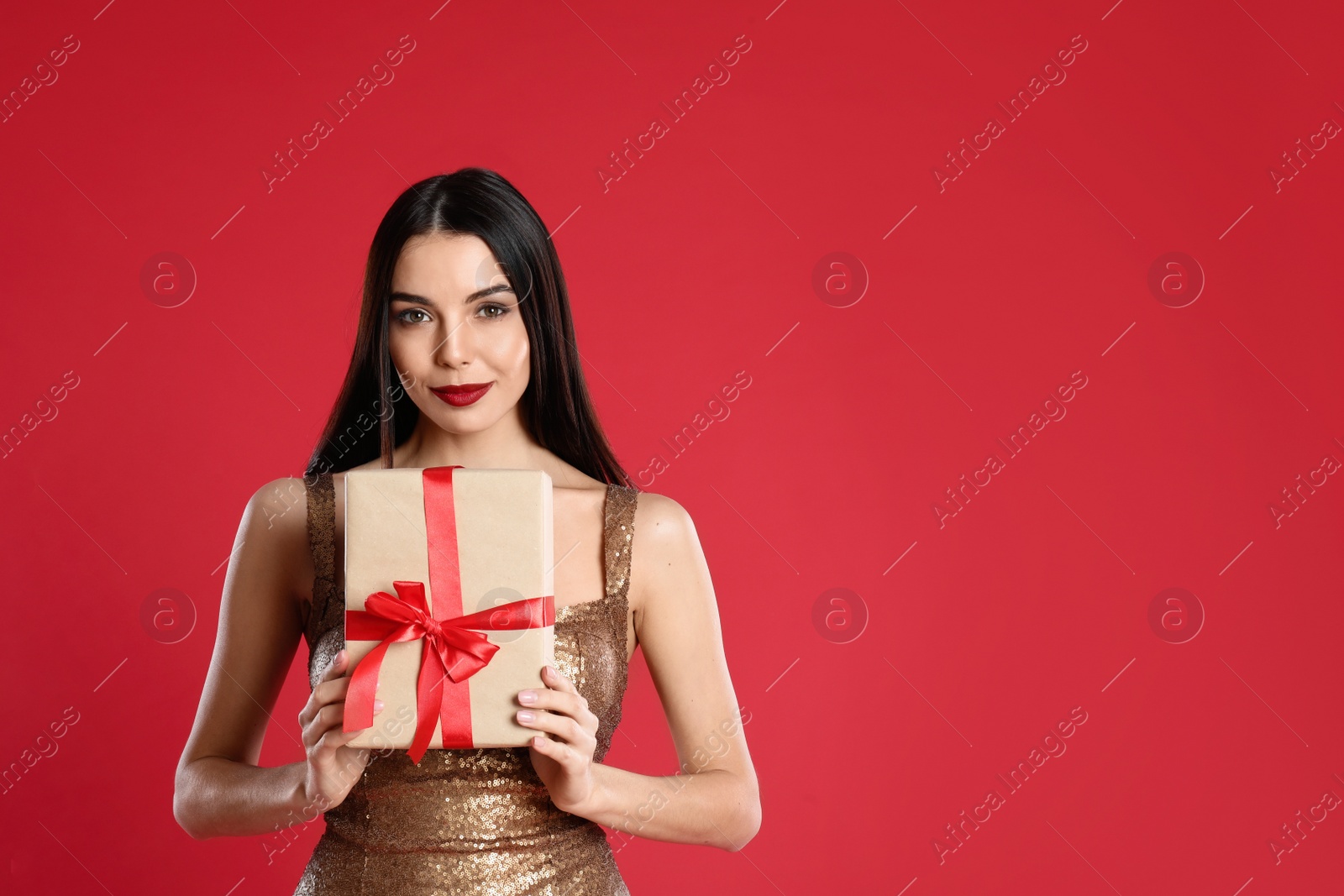 Photo of Woman in golden dress holding Christmas gift on red background, space for text