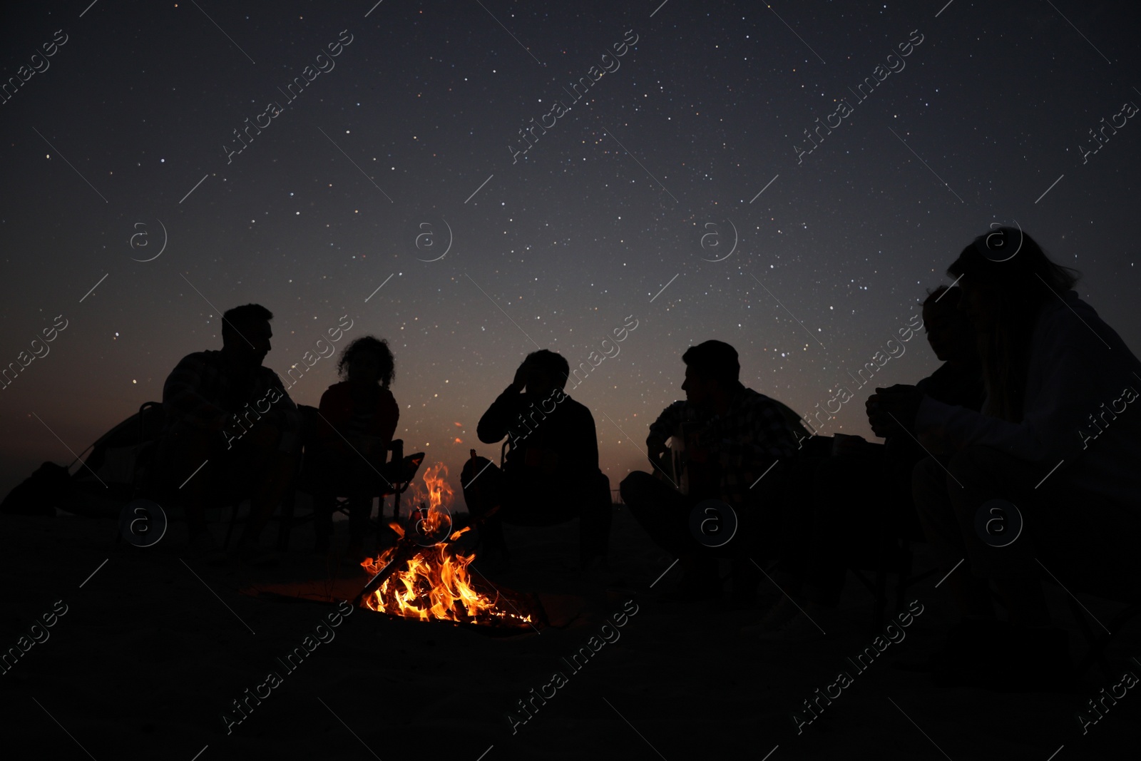 Photo of Group of friends gathering around bonfire in evening. Camping season