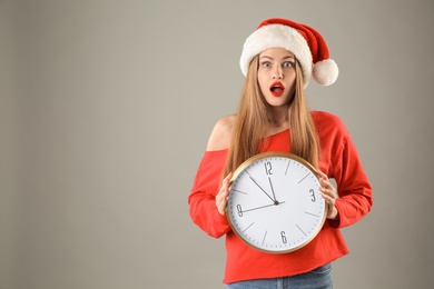 Young beautiful woman in Santa hat holding big clock on grey background. Christmas celebration