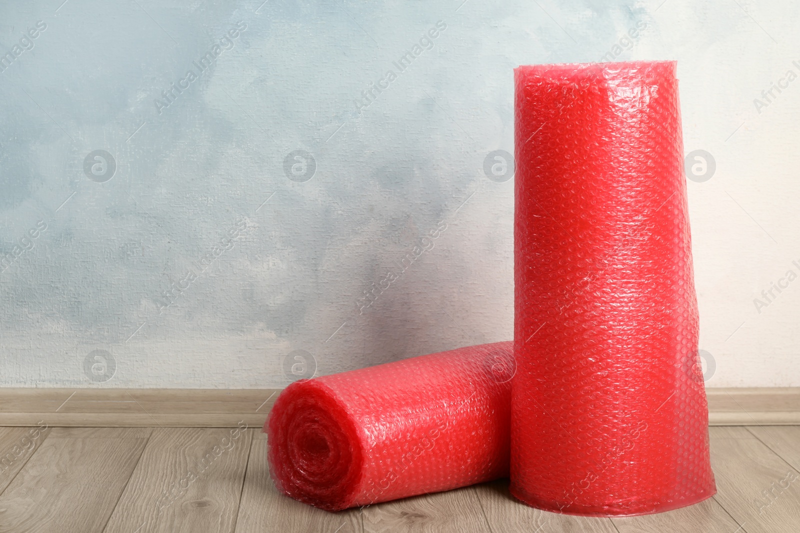 Photo of Red bubble wrap rolls on floor near light blue wall. Space for text