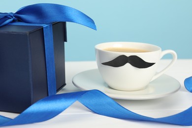 Happy Father's Day. Cup with moustache and gift box on white wooden table, closeup