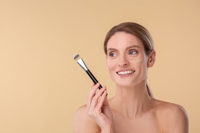 Woman with swatch of foundation holding makeup brush on beige background. Space for text