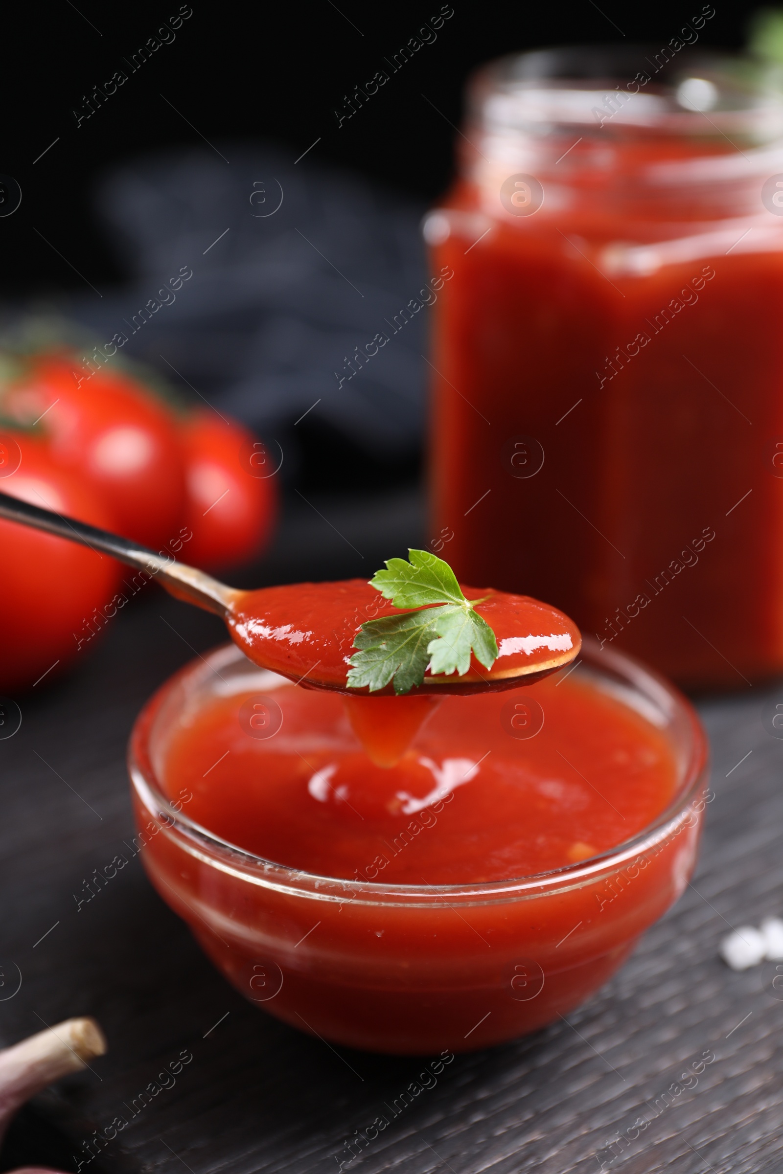 Photo of Taking tasty ketchup with spoon from bowl at black wooden table, closeup. Tomato sauce